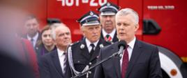 Minister Tomasz Siemoniak podczas Wojewódzkich Obchodów Dnia Strażaka. Szef MSWiA stoi przed mównicą, w tle strażacy i wóz strażacki. Na mównicy tabliczka z napisem: Państwowa Straż Pożarna, Komenda Wojewódzka PSP w Opolu.