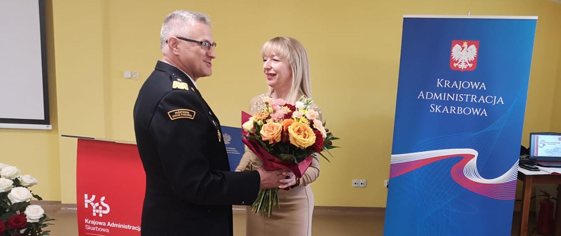 Z lewej Kujawsko-Pomorski Komendant Wojewódzki, obok niego Dyrektor. KAS. Komendant w granatowym mundurze wyjściowym wręcza Pani Dyrektor bukiet kwiatów. Po lewej w tle baner z napisem Krajowa Administracja Skarbowa.