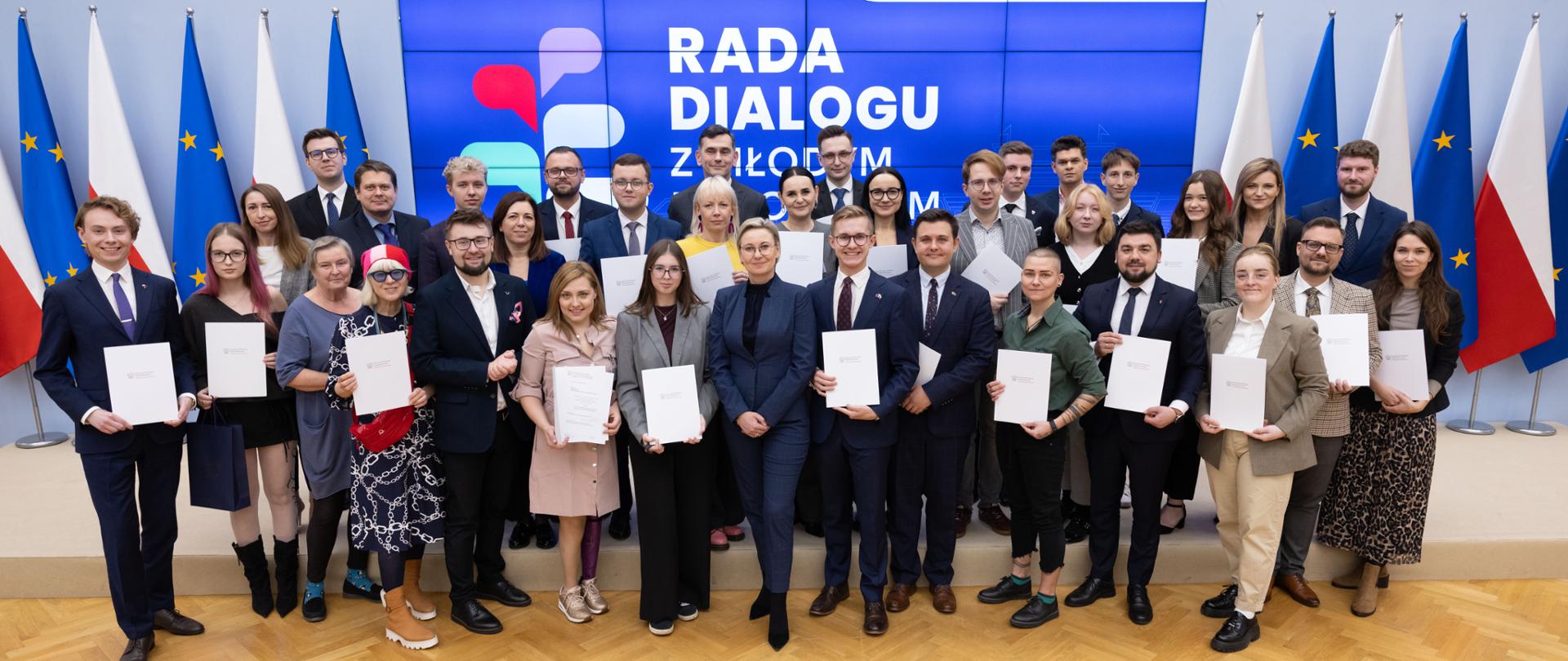 2025.10.30 Warszawa, Kancelaria Prezesa Rady Ministrów. I Posiedzenie Rady Dialogu z Młodym Pokoleniem IV kadencji. Zdjęcie grupowe na tle ekranu z logo i napisem Rada Dialogu z Młodym Pokoleniem. Na górze ekranu widoczne loga Kancelaria Prezesa rady Ministrów i Komitet do spraw Pożytku Publicznego. Po bokach ekranu widoczne są flagi Polski i Unii Europejskiej. fot. Stefan Maszewski