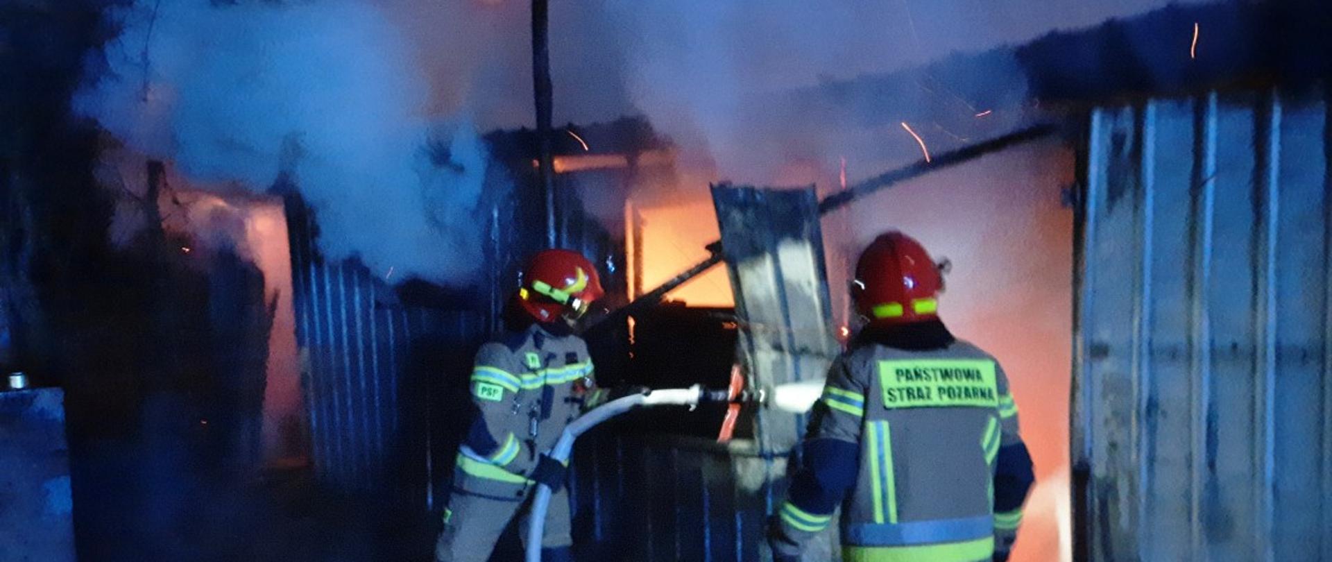 Zdjęcie zrobione jest w nocy i przedstawia dwóch strażaków gaszących palący się blaszany barak. Strażacy ubrani są w piaskowe stroje strażackie. Jeden ze strażaków trzyma w dłoni prądownicę z wężem i podaje strumień wody do wnętrza palącego się baraku. Obaj strażacy skierowani są twarzą w kierunku palącego się obiektu, a tyłem/bokiem do osoby wykonującej zdjęcie.