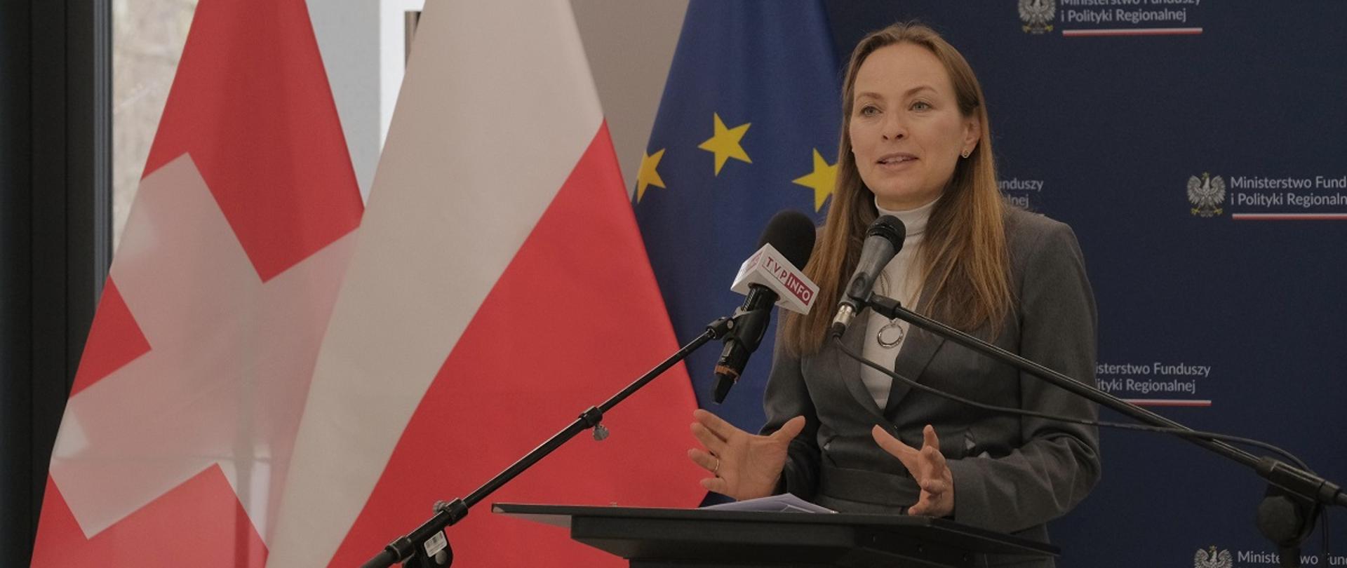 Minister funduszy i polityki regionalnej Katarzyna Pełczyńska-Nałęcz stoi przy mikrofonach, za jej plecami flagi Polski, Szwajcarii i UE