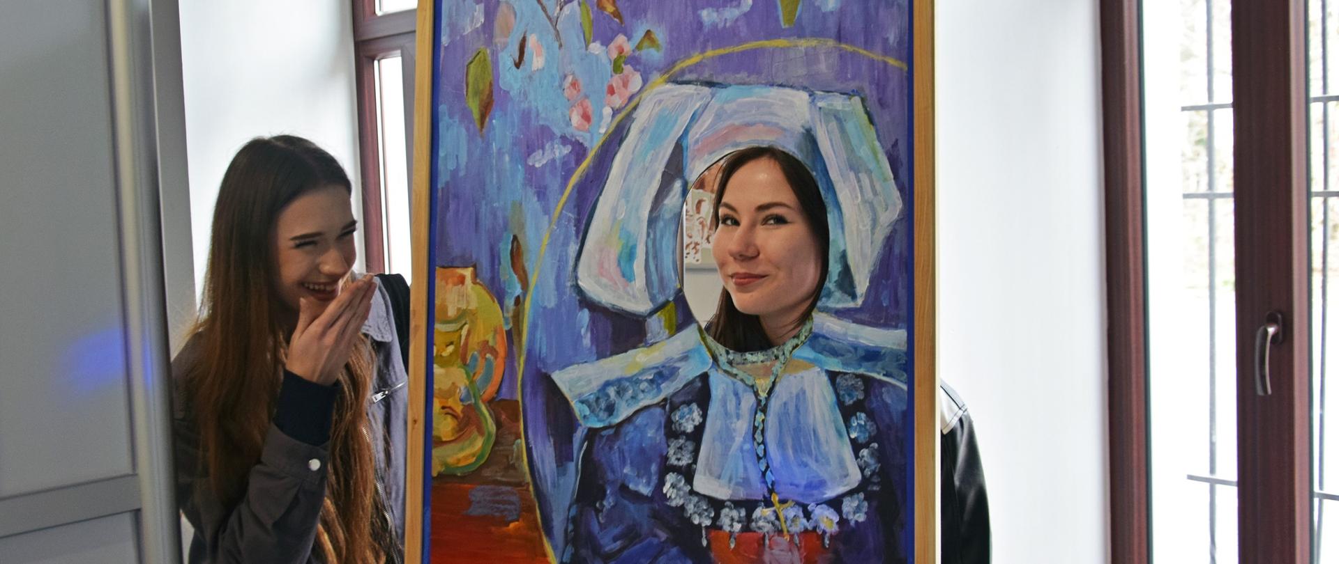 Obraz przedstawiający tradycyjny Strój francuski z czepcem, miejsce twarzy Wycięty otwór przez który widać twarz uśmiechniętej dziewczyny, obok obrazu stoi druga dziewczyna z długimi włosami, szeroko się uśmiecha przetrzymując rękę przy ustach.