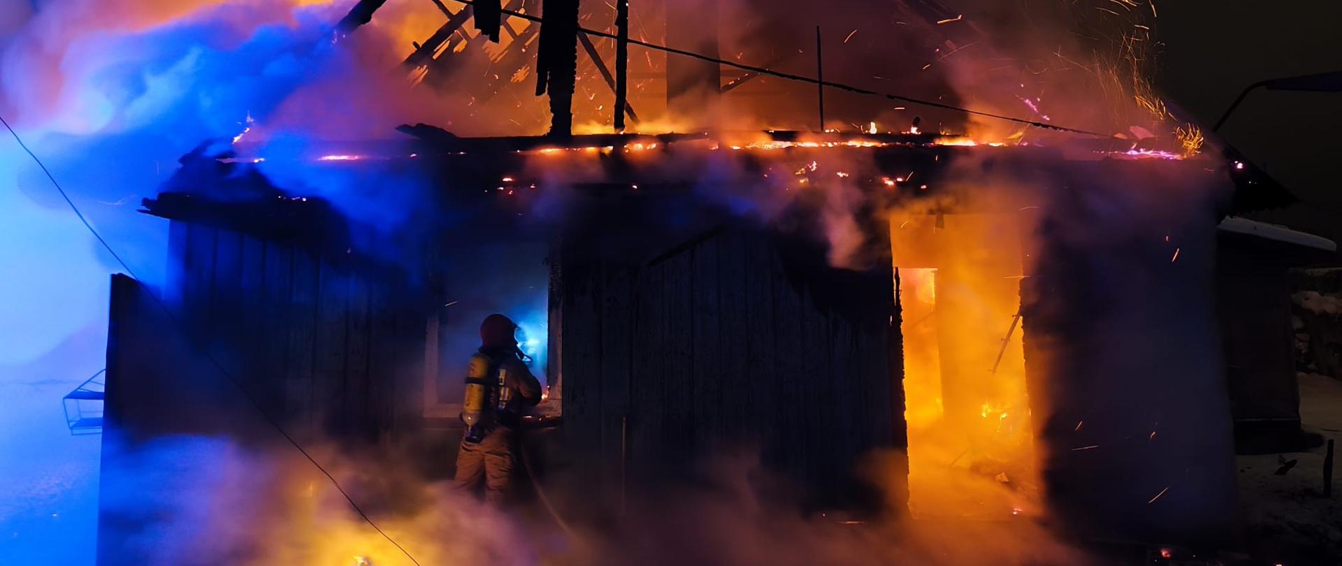 Pożar budynku w nocy. Strażak w kombinezonie ochronnym i z butlą powietrzną na plecach pracuje w pobliżu płonącej konstrukcji. Ogień jest intensywny, a gęsty dym rozświetlony jest przez światła awaryjne, nadając mu odcienie niebieskiego i fioletowego. Śnieg pokrywa ziemię, kontrastując z gorącem pożaru. Scena ukazuje dramatyzm i trudne warunki pracy służb ratowniczych.