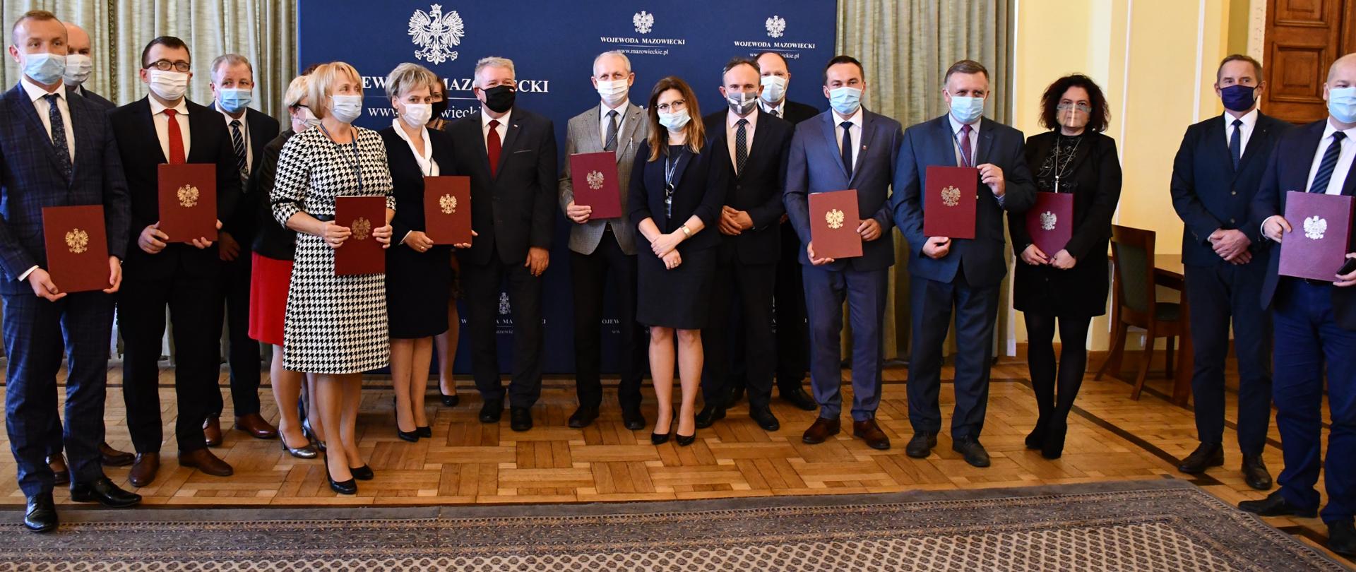 Na zdjęciu znajdują się elegancko ubrani przedstawiciele samorządów z województwa mazowieckiego z bordowymi teczkami w rękach oraz Wicewojewoda Mazowiecki. W tle znajduje się żółta ściana, zielone zasłony oraz granatowa ścianka z białymi napisami
