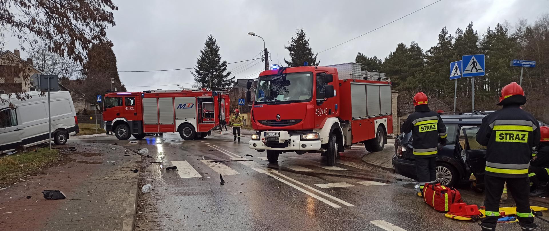 Na zdjęciu widać dwa auta biorące udział w zderzeniu w porze dziennej. Jeden rozbity samochód osobowy stoi po prawej strony jezdni, natomiast drugi po lewej stronie na poboczu. Tuż obok rozbitych samochodów stoją strażacy oraz zaparkowane dwa samochody ratowniczo-gaśnicze.