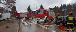 Na zdjęciu widać dwa auta biorące udział w zderzeniu w porze dziennej. Jeden rozbity samochód osobowy stoi po prawej strony jezdni, natomiast drugi po lewej stronie na poboczu. Tuż obok rozbitych samochodów stoją strażacy oraz zaparkowane dwa samochody ratowniczo-gaśnicze.