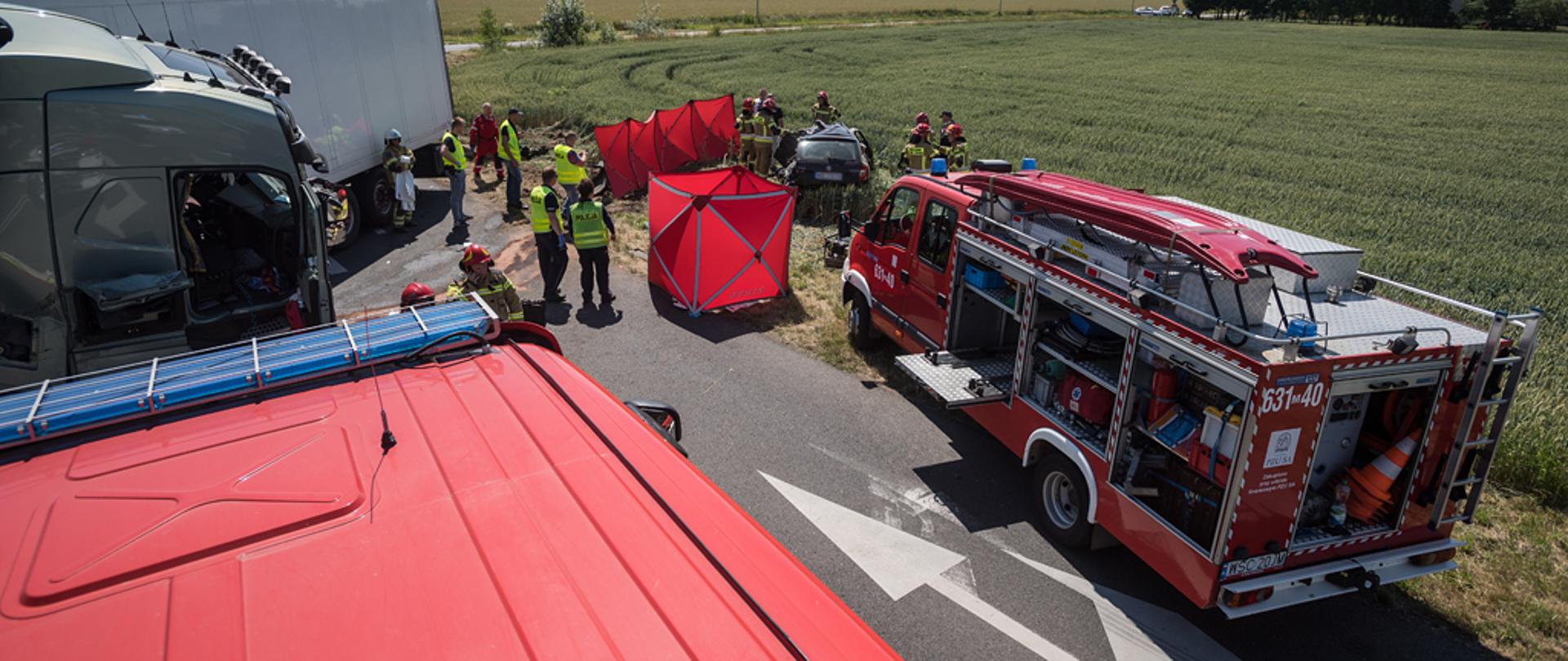 Zdjęcie wykonane z dachu samochodu pożarniczego przedstawia ogólny widok na miejsce zdarzenia. Widoczny rozbity samochód osobowy oraz fragment ciągnika siodłowego z naczepą. Na miejscu pracują strażacy i policjanci.
