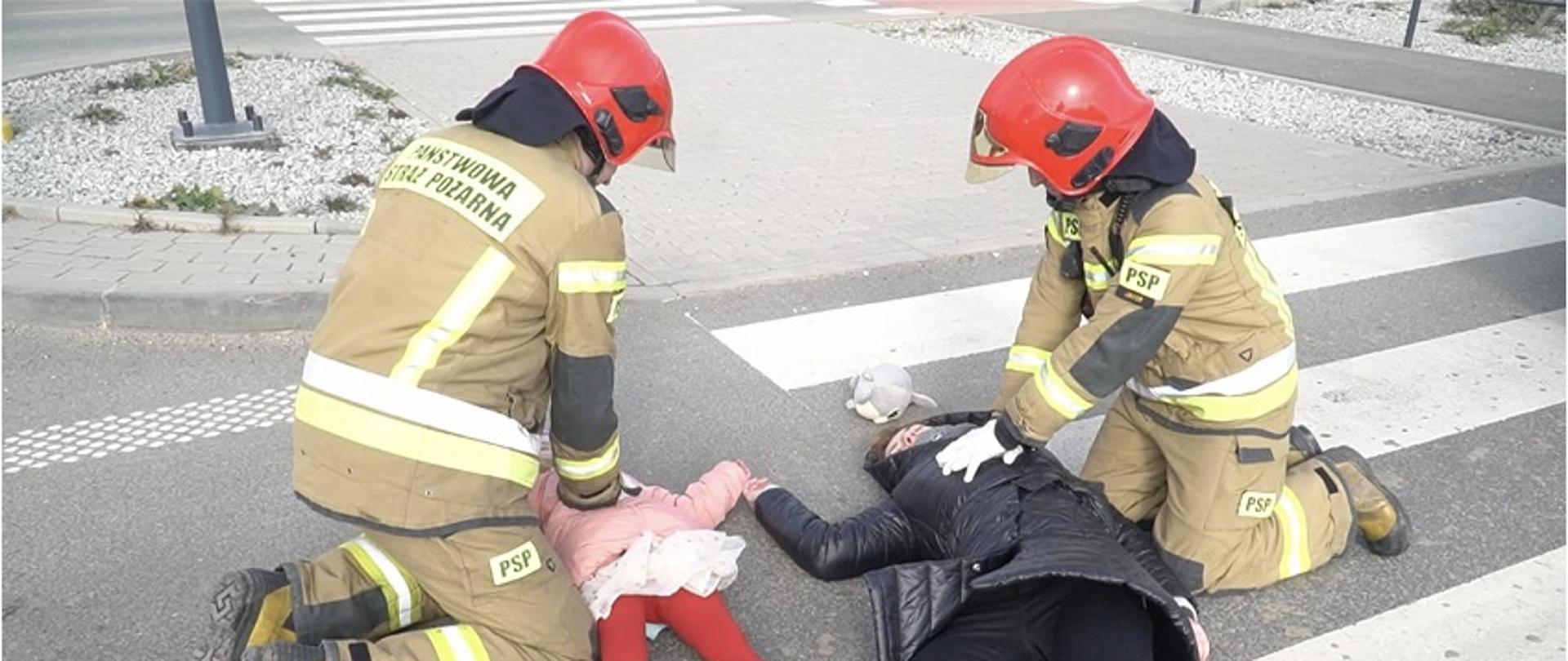 Obraz przedstawia strażaków prowadzących resuscytację krążeniowo-oddechową kobiety i dziecka, którzy zostali potrąceni na pasach.