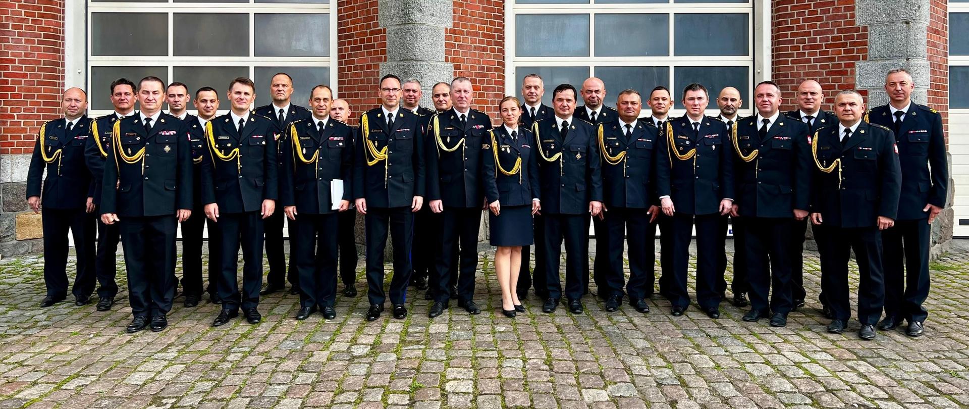 Zdjęcie przedstawia dużą grupę strażaków, stojących przed budynkiem. Jest to frontowe ujęcie całej grupy. Mężczyźni i kobieta stoją w rzędzie, z rękami ułożonymi wzdłuż tułowia, w postawie na baczność.
Wszyscy ubrani są w granatowe, galowe mundury strażackie, ozdobione złotymi guzikami i naszywkami. Mundury posiadają również białe kołnierzyki koszul i złote dystynkcje na rękawach. Kobieta w grupie, stojąca w środku, jest ubrana w taką samą kurtkę mundurową i granatową spódnicę. Wszyscy wyglądają na zadowolonych i uśmiechniętych.
Grupa stoi na brukowanym podłożu. W tle znajduje się czerwony, ceglany budynek z dwoma dużymi bramami wjazdowymi, które są zamknięte i mają numery "4" i "3" nad nimi. Nad bramami widać duże, prostokątne okna z podziałami. Dach budynku jest ciemny, a u góry widać ozdobne wykończenie. Całe ujęcie jest ostre i dobrze oświetlone.
