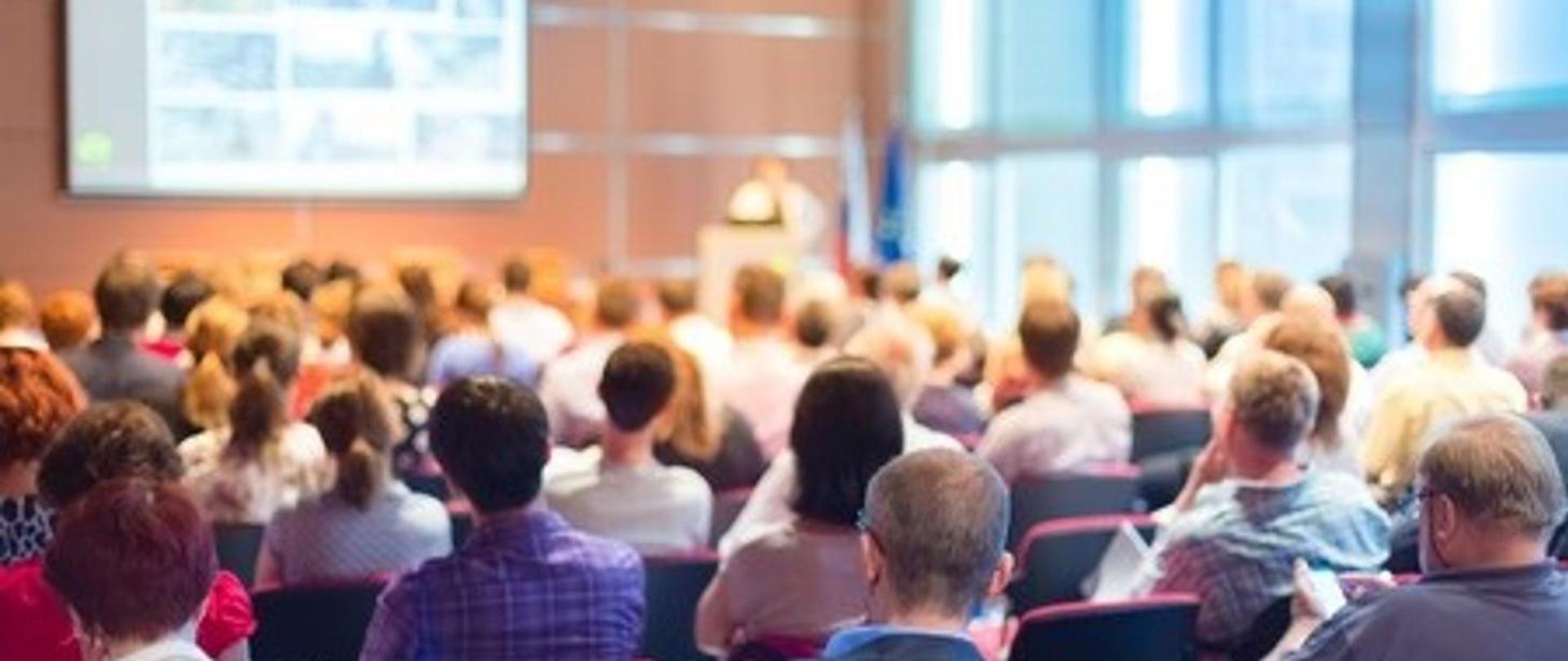 Na sali wykładowej duży ekran. Ludzie siedzą na krzesłach