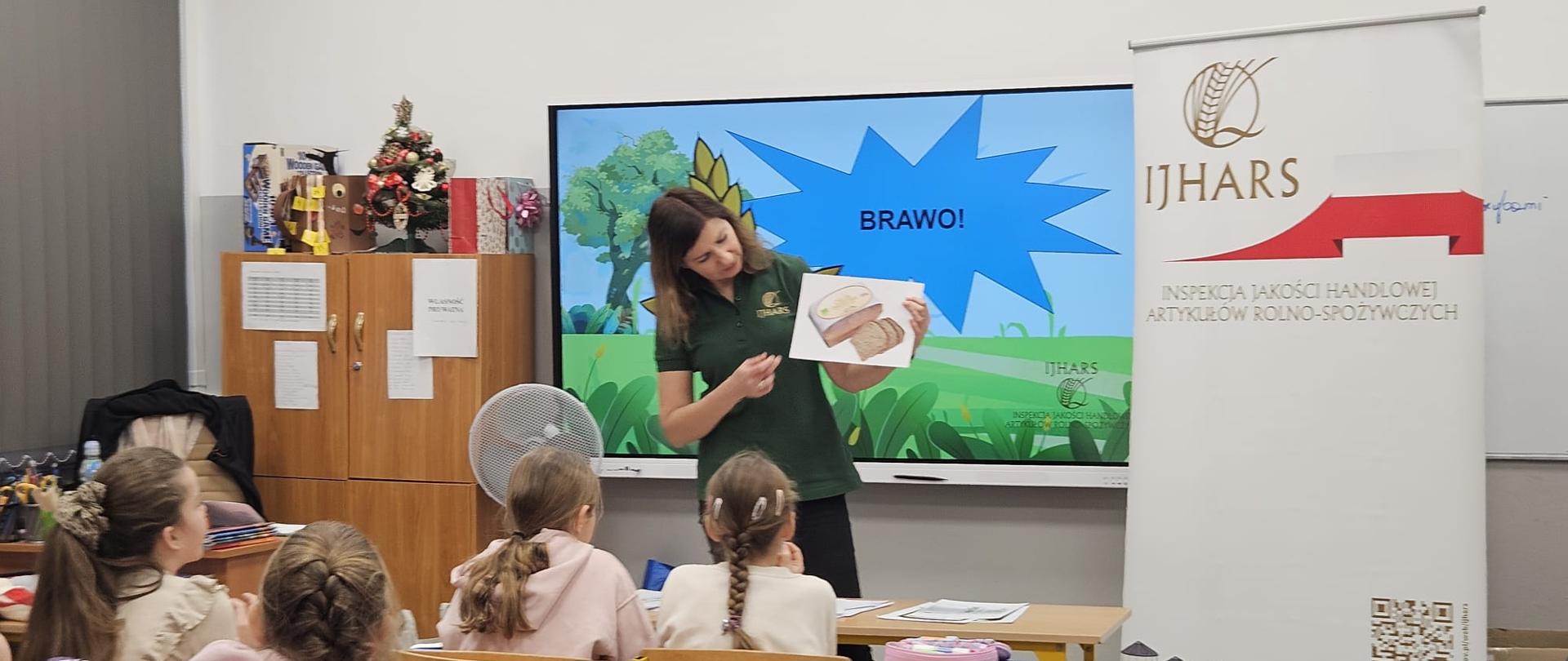 Inspektorka IJHARS prowadzi lekcję w Szkole Podstawowej nr 51 – stoi przy tablicy, a w tle widoczny jest roll-up z logo IJHARS; uczniowie siedzą w ławkach i uważnie słuchają.