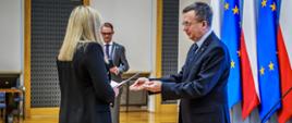 Szef służby cywilnej Dobrosław Dowiat-Urbański wręcza akt mianowania nowej urzędniczce służby cywilnej - w tle flagi Polski i Unii Europejskiej