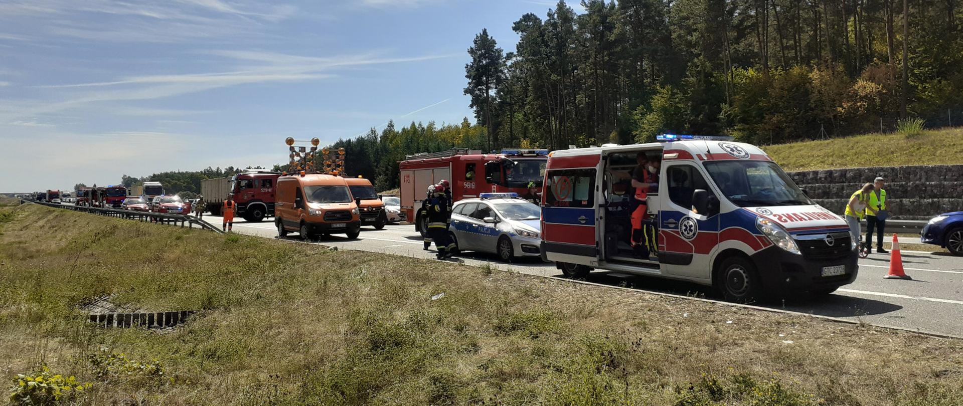 Zdjęcie przedstawia widok miejsca zdarzenia. Widać służby ratownicze podczas działań, służby autostradowe.
