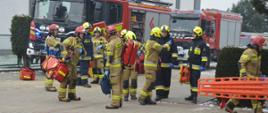 Plac przed budynkiem szkoły. Na tle hali sportowej stoją trzy wozy strażackie. Przed nimi strażacy w ubraniach specjalnych w kolorach ciemnych i piaskowych oraz w czerwonych i żółtych hełmach. Część z nich ma ze sobą torby ze sprzętem ratownictwa medycznego oraz nosze typu deska. 