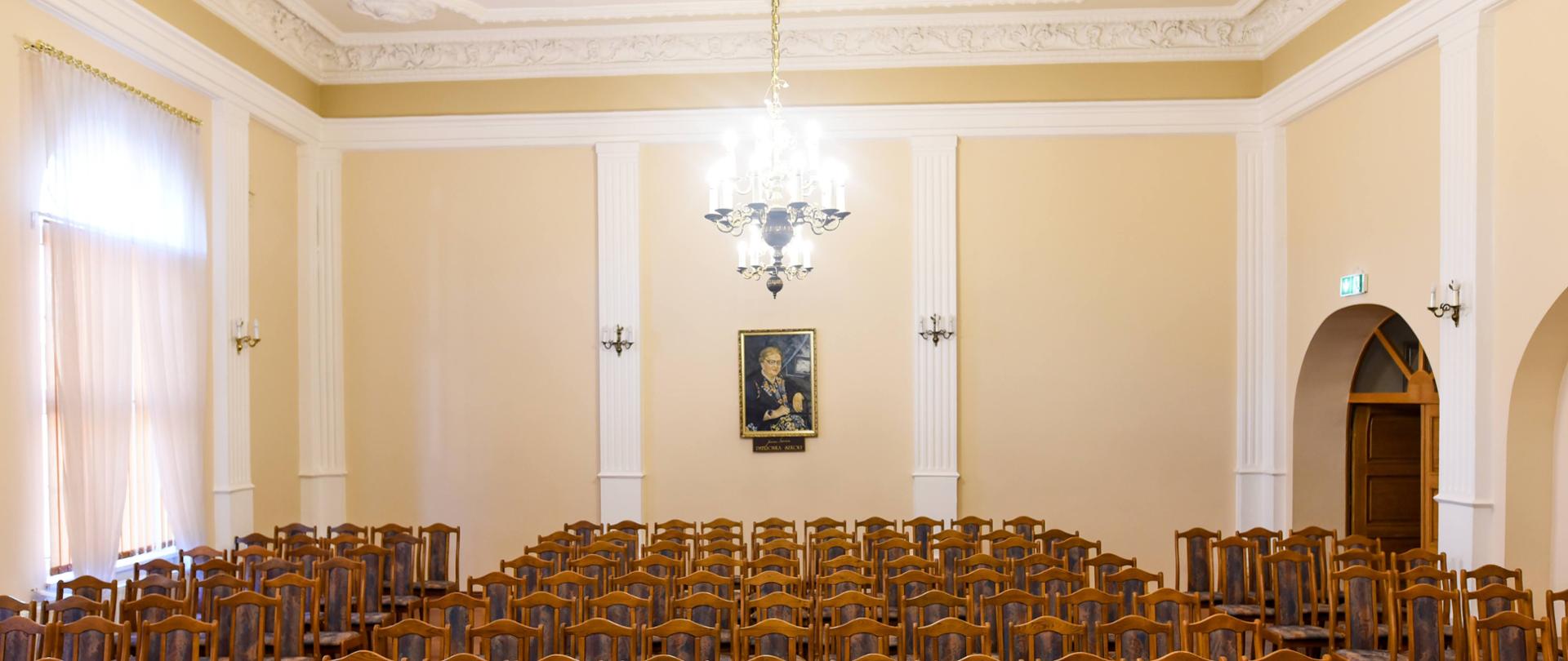 sala koncertowa Państwowej Szkoły Muzycznej I stopnia imienia Janiny Garści w Lublińcu - widok ze sceny na widownię: jasnobrązowe tapicerowane krzesła ustanowione w rzędach, ściany jasnożółte z białymi obwódkami, biały sufit ze sztukaterią, oświetlony żyrandol, na środku tylnej ściany portret patronki szkoły