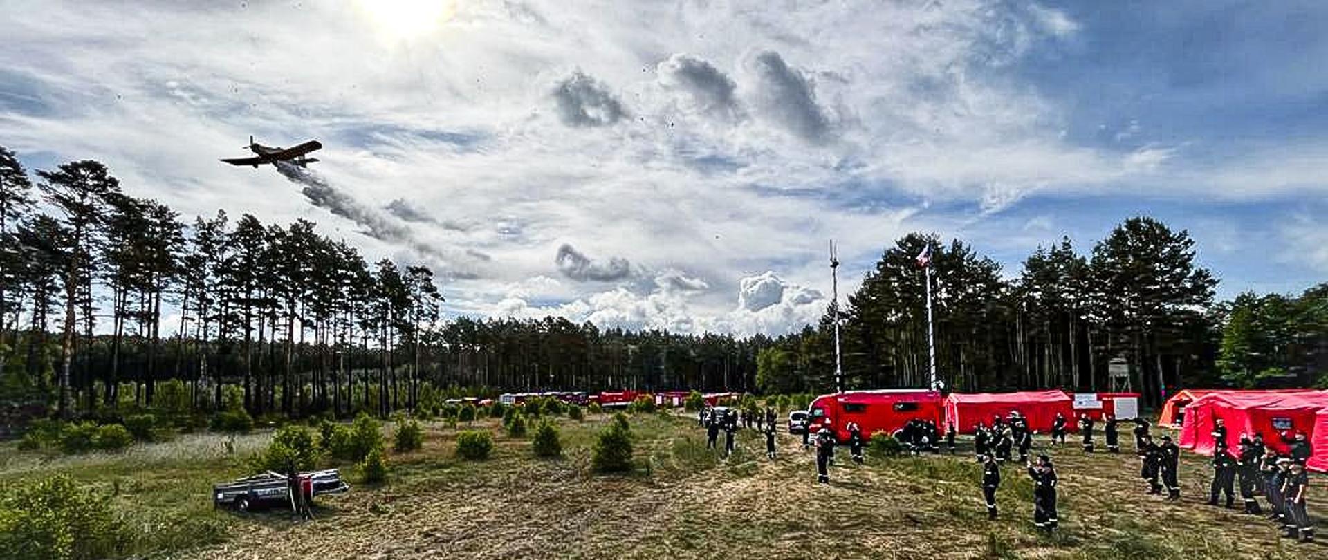 Zdjęcie przedstawia strażaków obserwujących zrzut wody wykonywany przez samolot gaśniczy. Po prawej stronie widać miasteczko namiotowe i samochody batalionowe. W tle las i samochody gaśnicze.