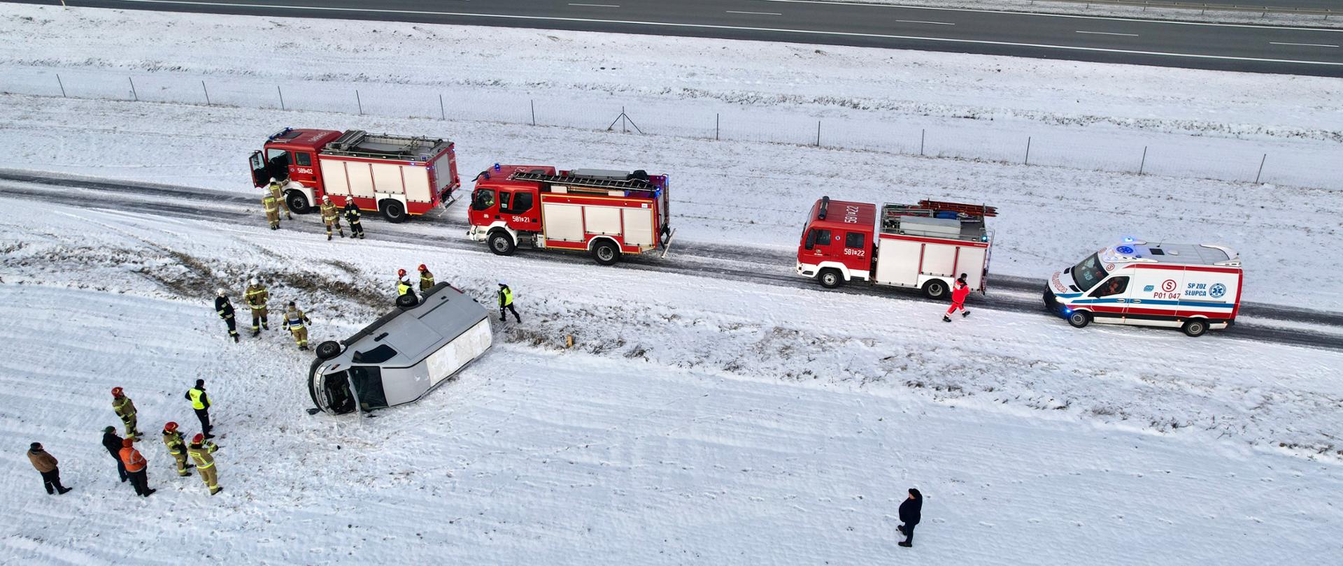 Biały samochód typy BUS lezy na boku poza droga wzdłuż autostrady wokół niego strażacy na drodze stoją trzy samochody pożarnicze oraz jeden samochód karetki pogotowia w oddali widać odcinek autostrady.