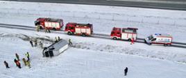Biały samochód typy BUS lezy na boku poza droga wzdłuż autostrady wokół niego strażacy na drodze stoją trzy samochody pożarnicze oraz jeden samochód karetki pogotowia w oddali widać odcinek autostrady.