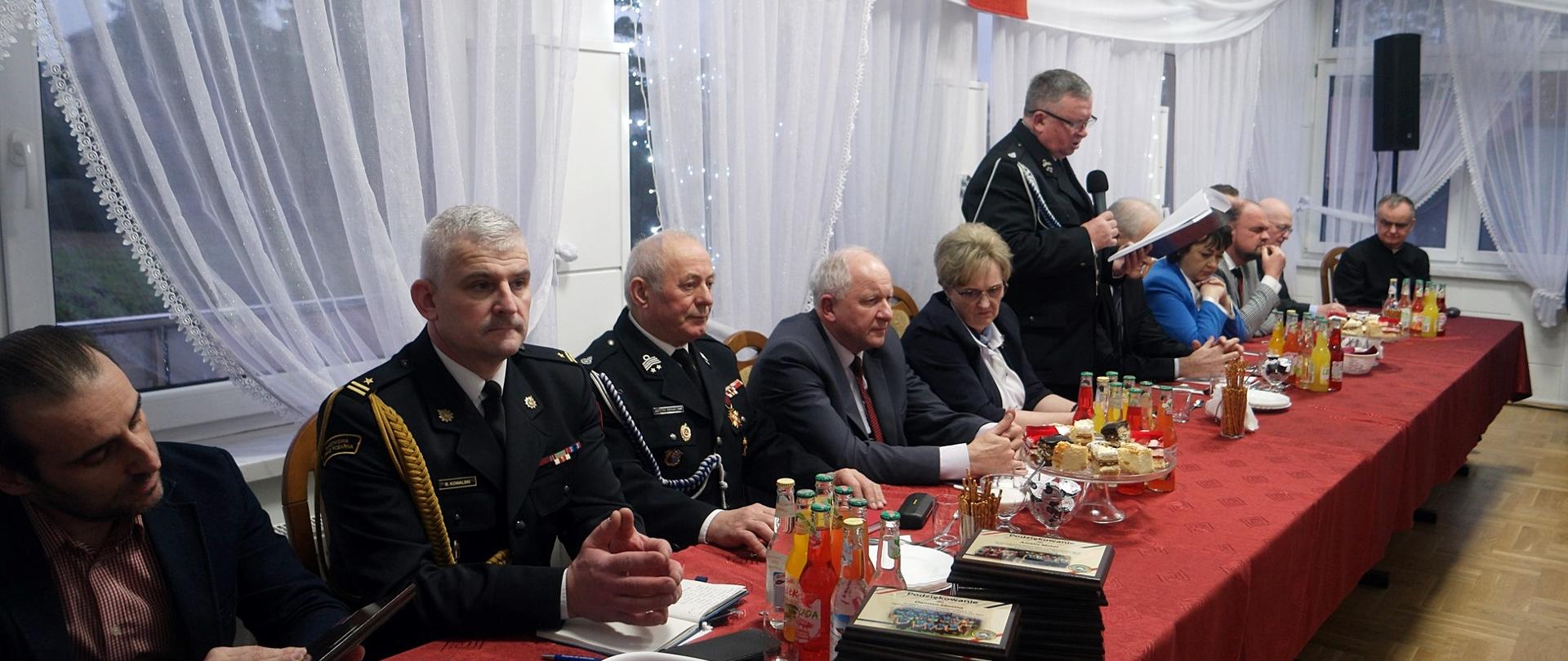 Widok boczny, zdjęcie zbiorowe zaproszonych gości siedzących przy wspólnym stole. Jedna osoba stoi i przemawia do mikrofonu.