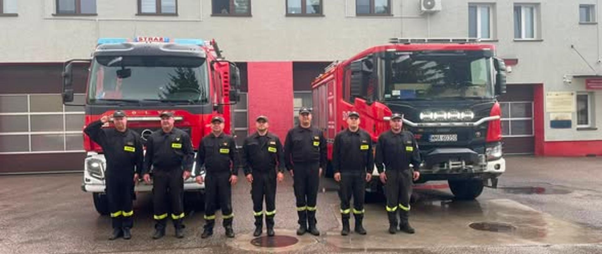 Na zdjęciu widać siedmiu funkcjonariuszy PSP, którzy oddają hołd z okazji 82. rocznicy wybuchu Powstania Warszawskiego. Za nimi stoją dwa wozy strażackie. Z tyłu widać budynek Komendy. 