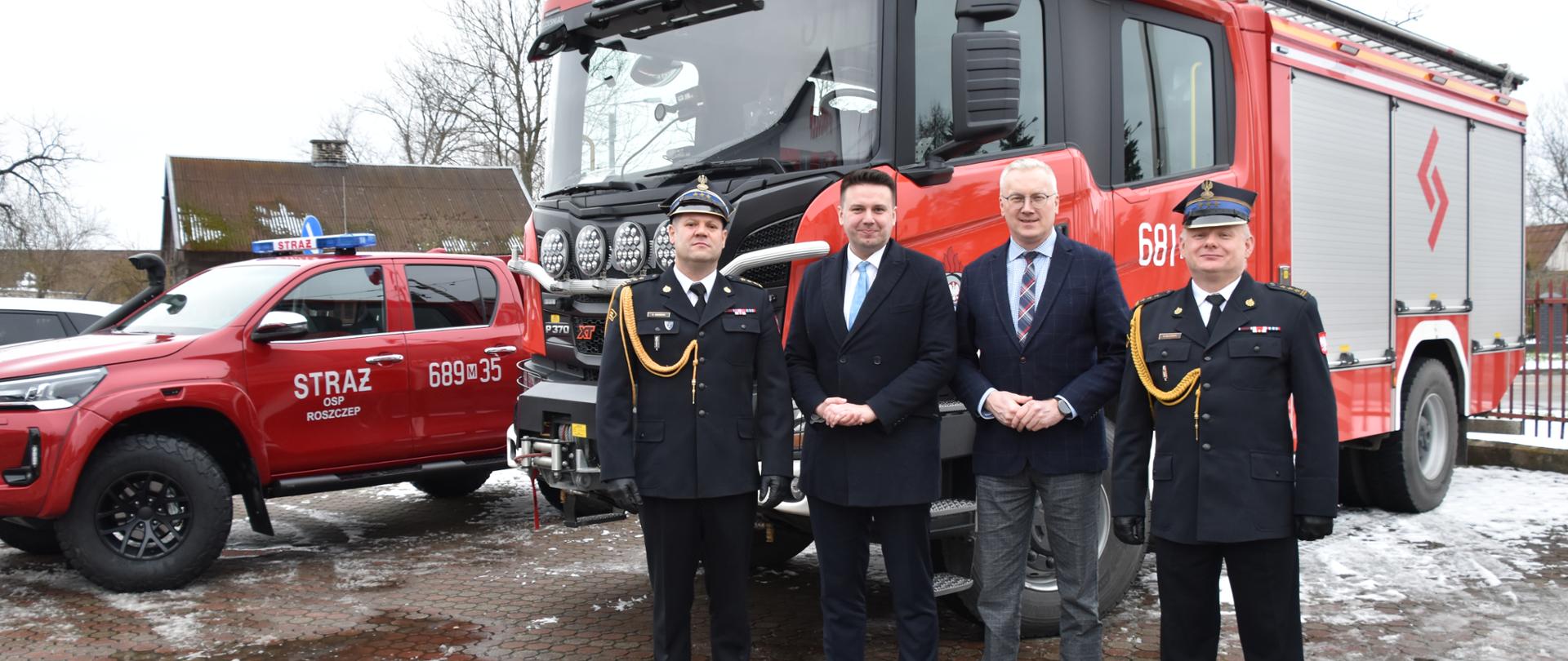 Zdjęcie przedstawia stojących przed samochodami strażackimi mężczyzn, dwóch ubranych jest w garnitury, natomiast dwóch w mundury strażackie. Strażacy mają czapki na głowach. Na boku jednego z pojazdów widnieje napis OSP Roszczep