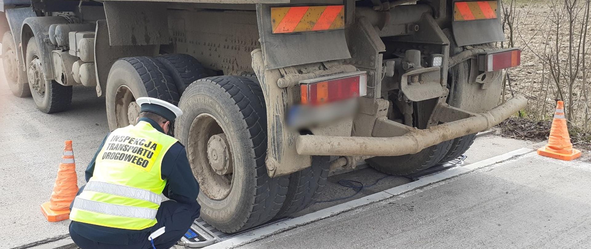 Od lewej: umundurowany inspektor warmińsko-mazurskiej Inspekcji Transportu Drogowego odczytuje wynik pomiaru z wag inspekcyjnych, przez które przejeżdża wywrotka. Samochód ciężarowy stoi kołami ostatniej osi na wagach.