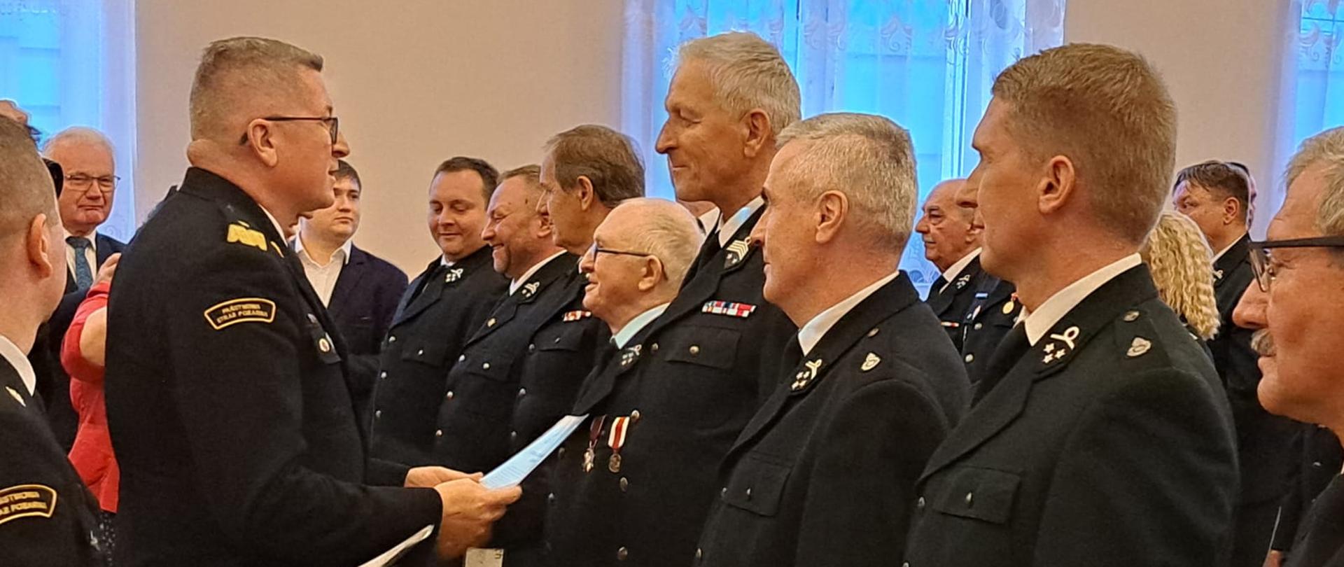 Zastępca komendanta głównego wręcza dokument druhowi który soi w rzędzie z razem z innymi przedstawicielami jednostek OSP . Wszyscy ubrani są w mundury strażackie