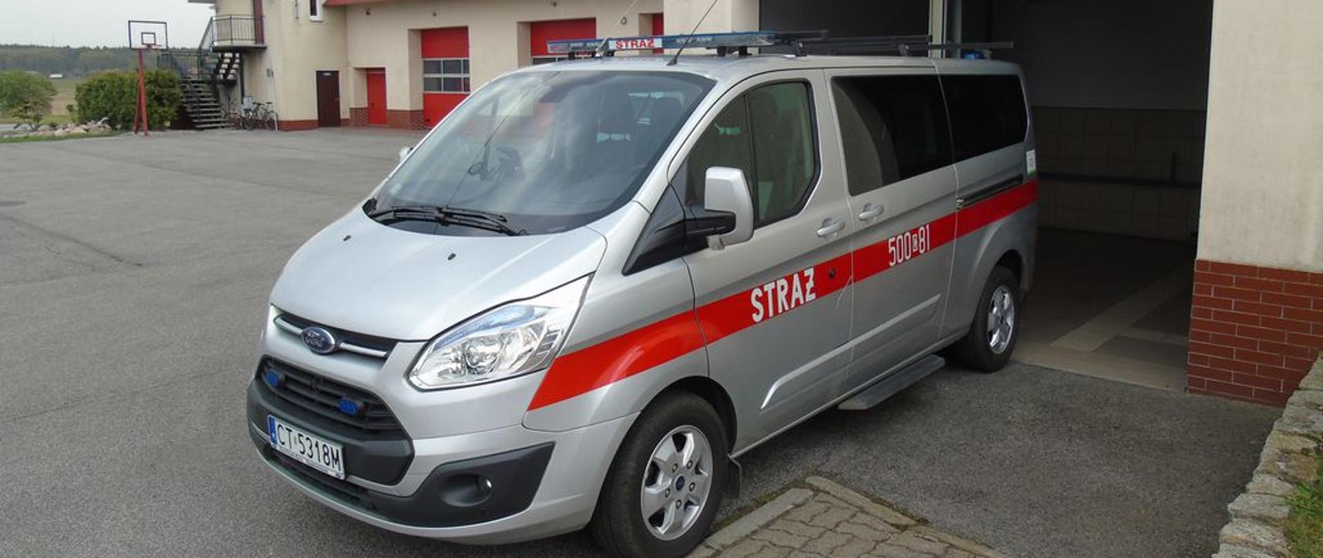 Zdjęcie przedstawia srebrny samochód SLKW Ford bus na tle garażu KP PSP.