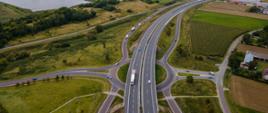 Widok z lotu ptaka na dwujezdniową drogę ekspresową z węzłem drogowym. Jezdnie rozdzielone pasem zieleni, po bokach drogi serwisowe i pola uprawne. W tle widoczne zbiorniki wodne, zabudowa miejscowości oraz rozległy krajobraz rolniczy pod pochmurnym niebem.
