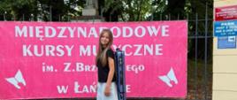 Młoda dziewczyna z długimi, jasnymi włosami stojąca na tle różowej tablicy informacyjnej z napisem "MIĘDZYNARODOWE KURSY MUZYCZNE im. Z. Brzewskiego w Łańcucie". Dziewczyna ma na sobie czarną koszulkę, jasne, luźne spodnie oraz białe buty, a na plecach niesie futerał na skrzypce. W tle widać zabytkową bramę i drzewa.
