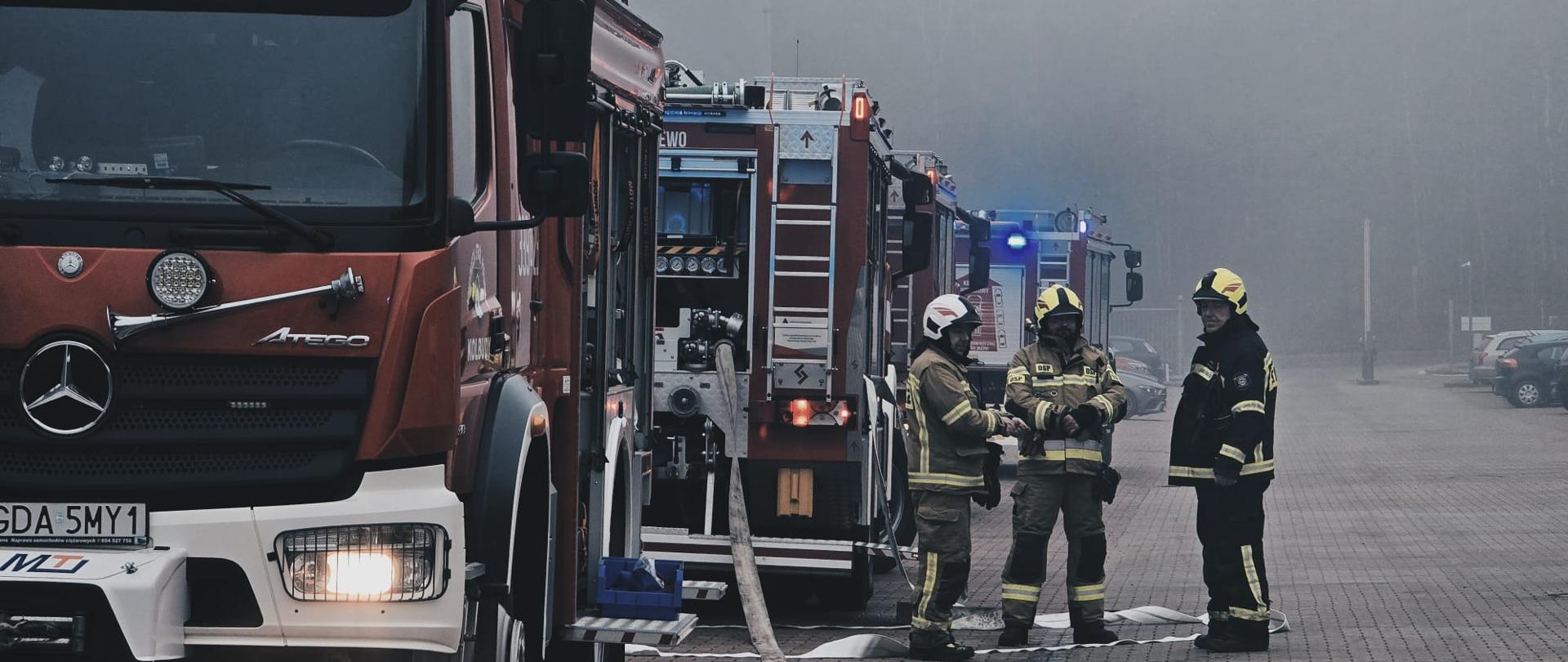 Zdjęcie przedstawia wozy bojowe i strażaków podających wodę do pożaru