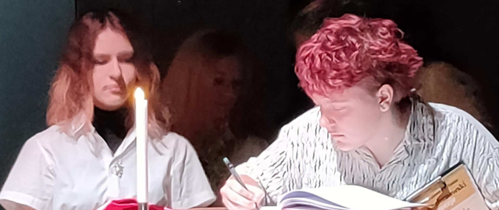 Dwie uczennice siedzące przy stole, na którym stoi świeca, zdjęcie w półmroku. 