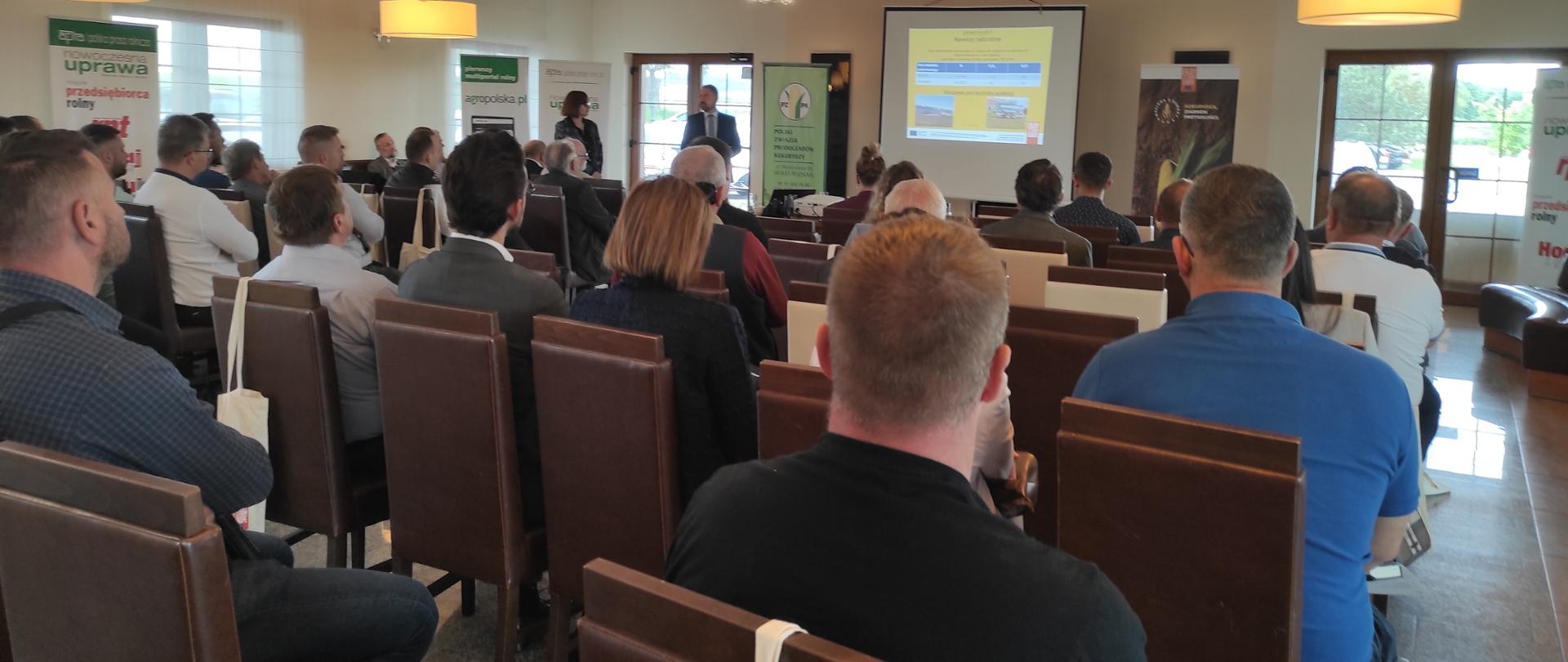 Osoby siedzące na sali konferencyjnej z wykładowcą. 