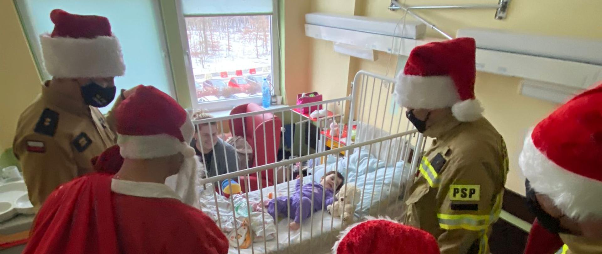 Strażacy w mundurach w sali szpitalnej przy łóżeczku dziecka.