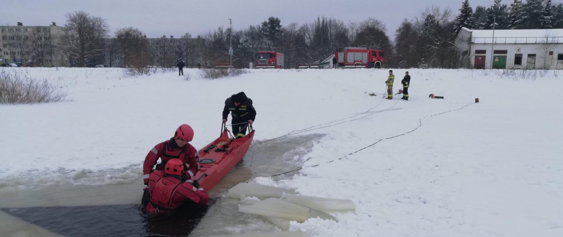 Na zamarzniętym akwenie znajduje się przerębel, z którego dwóch strażaków przy użyciu "sań lodowych" wyciąga osobę znajdującą się w wodzie. Na brzegu stoi dwóch strażaków trzymających linki podpięte do "sań lodowych", widoczne są również samochody pożarnicze oraz zabudowania. 