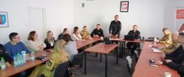 W sali konferencyjnej w siedzibie KP PSP w Ostrowcu Św. siedzą strażacy oraz pracownicy cywilni przy stolikach i słuchają wykładu. Na stojąco Zastępca KP PSP w Ostrowcu Św. objaśnia zgromadzonym na sali zagadnienia.