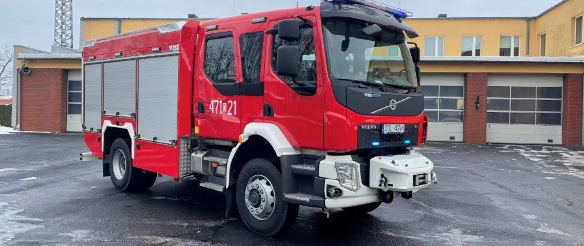 Samochód pożarniczy koloru czerwonego stoi na placu, z tył widać budynek strażnicy żółty z bramami garażowymi koloru białego.