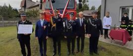 Na pierwszym planie osoby przekazujące pojazd pożarniczy ustawione w szeregu, wśród nich Poseł na Sejm, przedstawiciele samorządu, komendant wojewódzki PSP oraz druhowie OSP, za nimi przekazywany pojazd