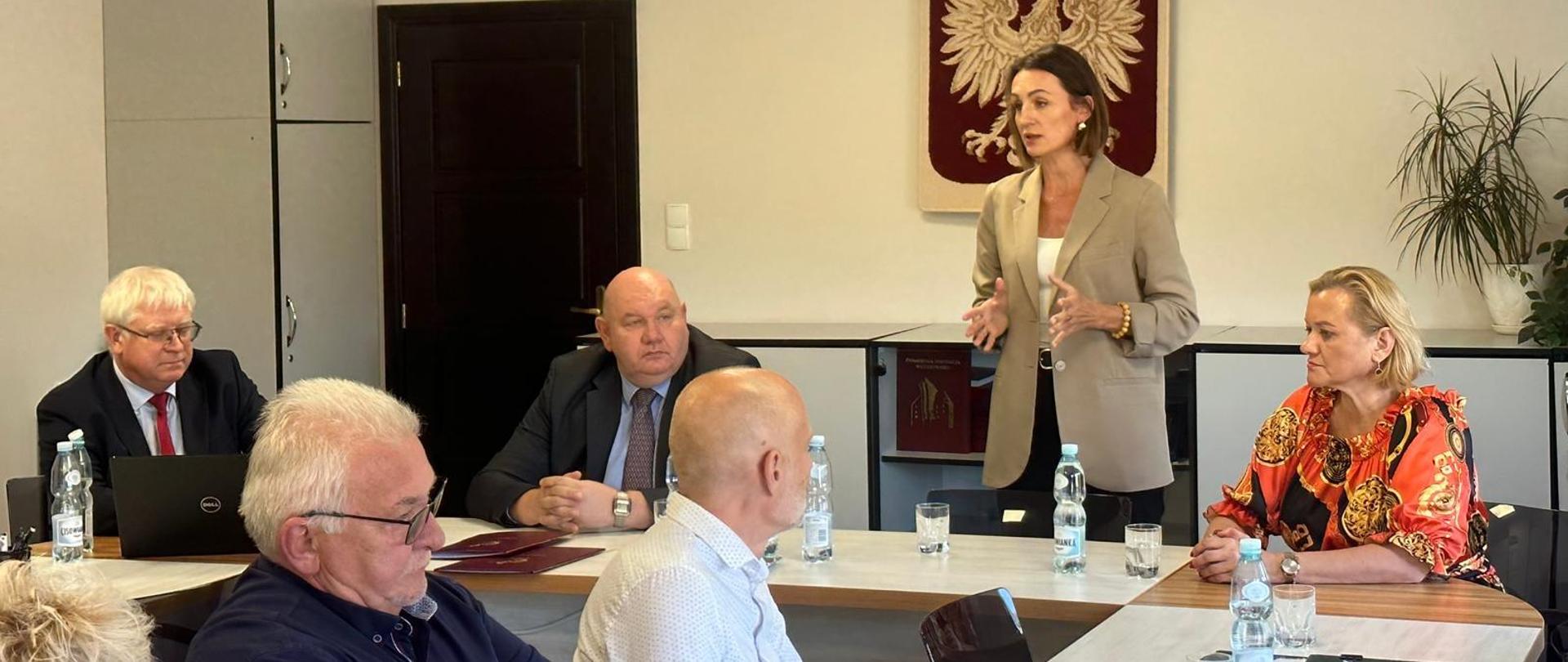 Kilka osób siedzi przy dużym stole. Kobieta (wojewoda) stoi i przemawia