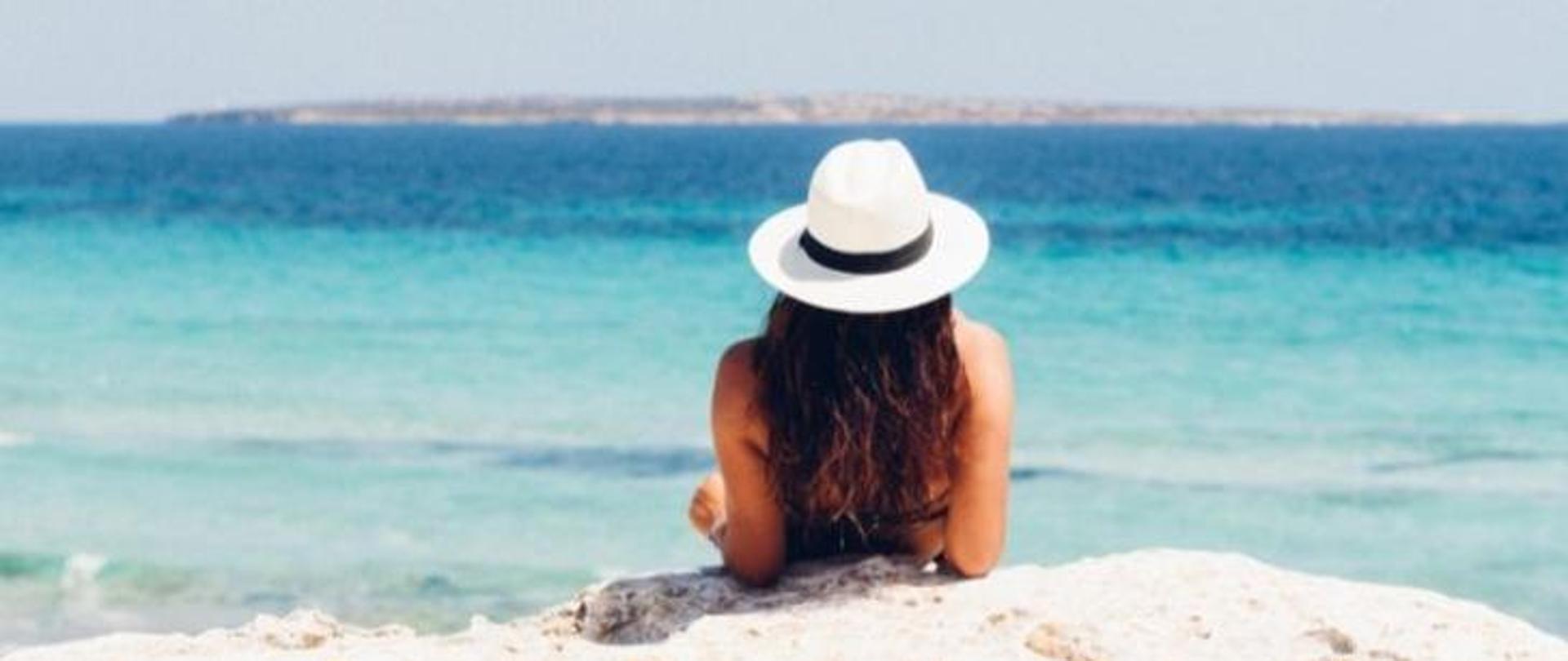 Brunetka mająca na głowie biały kapelusz wypoczywa na brzegu morza