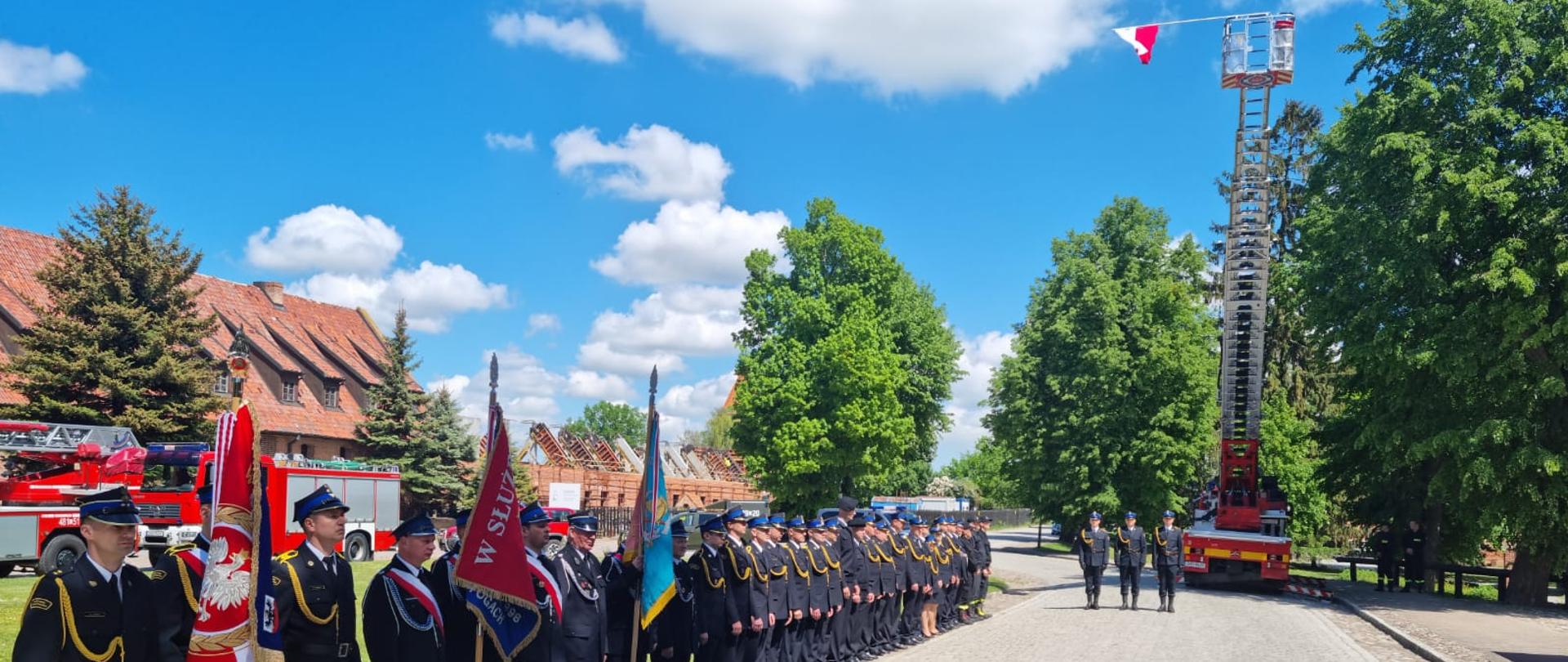 Fotografia przedstawia strażaków stojących na apelu z okazji Dnia Strażaka oraz samochód pożarniczy z drabiną mechaniczną, który podnosi flagę państwową.