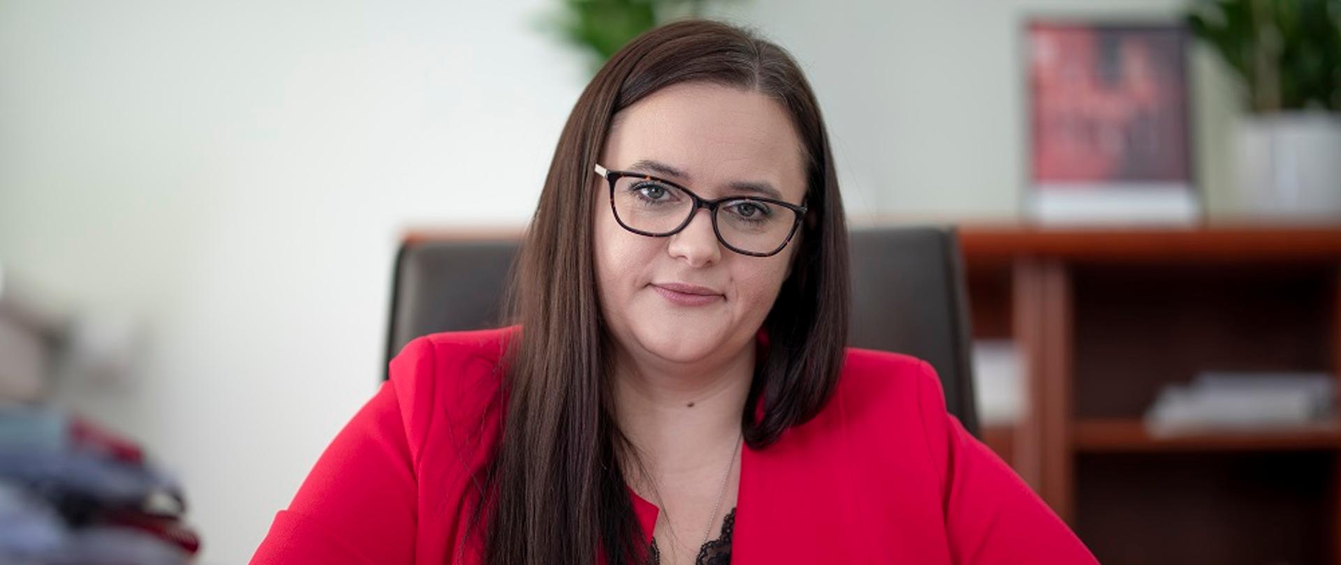 Portret minister Jarosińskiej-Jedynak, w różowym żakiecie. Pani Minister siedzi przy biurku i podpisuje dokumenty