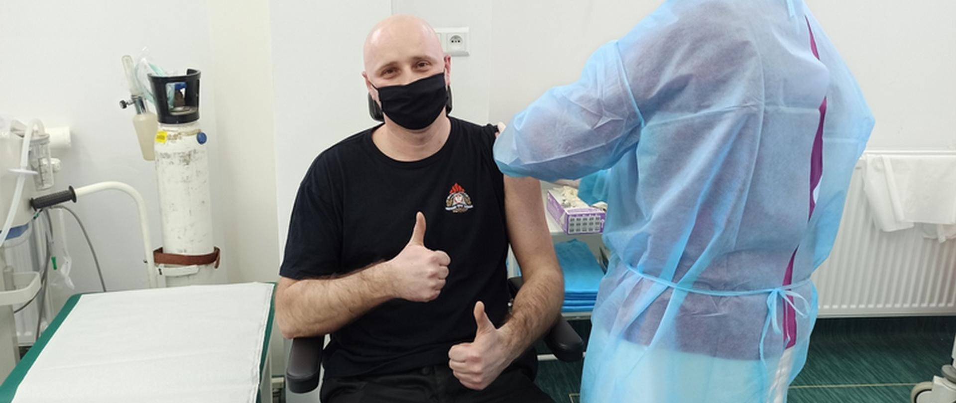 Zdjęcie przedstawia strażaka, którego personel medyczny szczepi przeciwko COVID-19. Straż pokazuje wyciągnięty kciuk na znak akceptacji i pozytywnego podejścia do szczepienia.