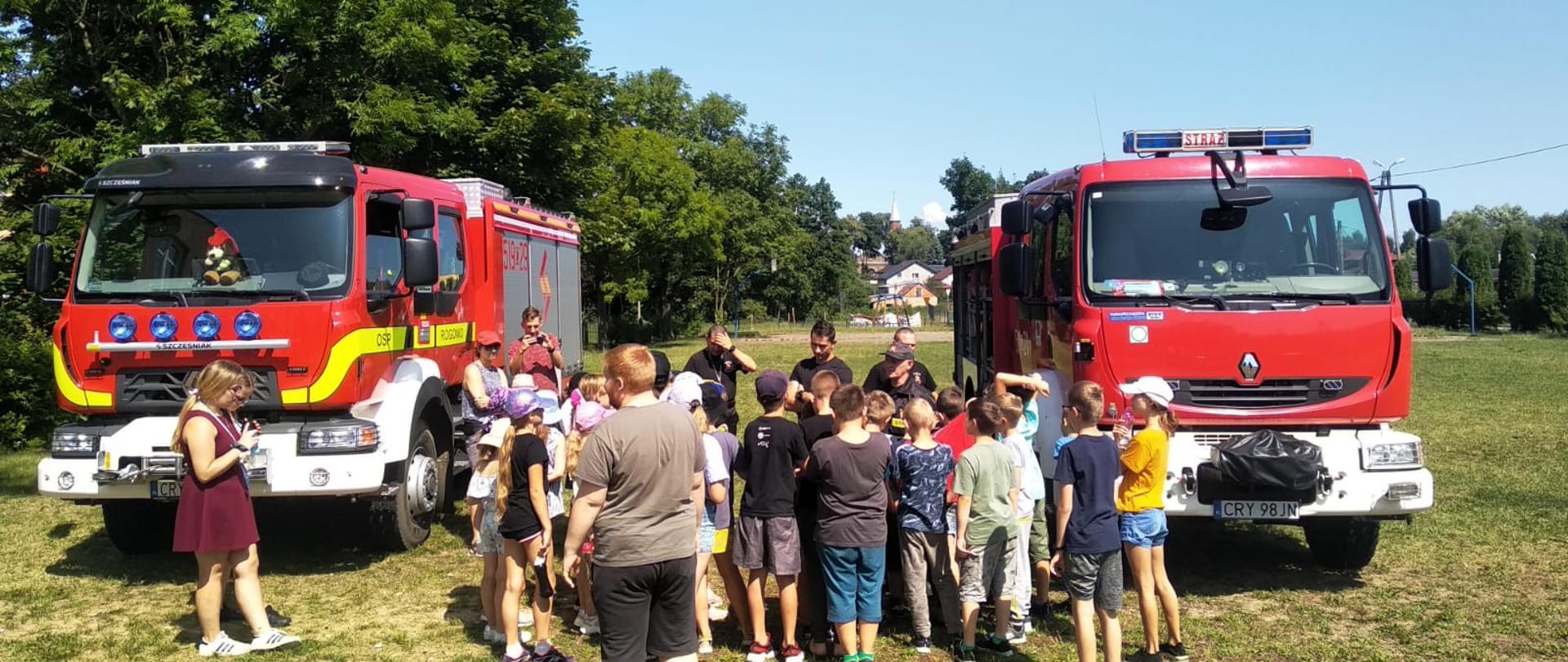 Zdjęcie wykonane jest na boisku i przedstawia grupę młodzieży, która słucha strażaków. Po lewej i prawej stronie zdjęcia stoi wóz strażacki. W tle zdjęcia widać drzewa oraz budynki. 