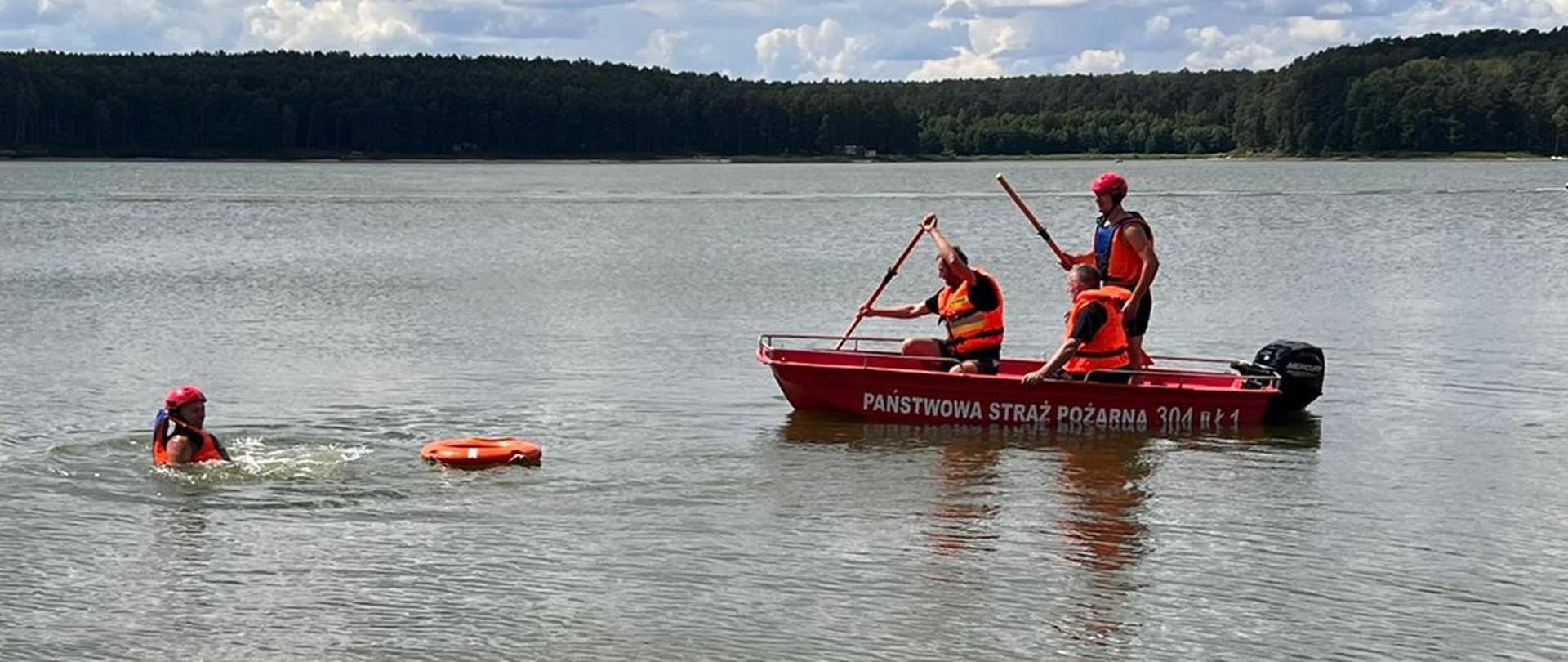 Zdjęcie przedstawia trzech strażaków w łodzi ratowniczej, którzy podpływają do zabezpieczonego w kamizelkę ratunkową funkcjonariusza udającego osobę poszkodowaną 
