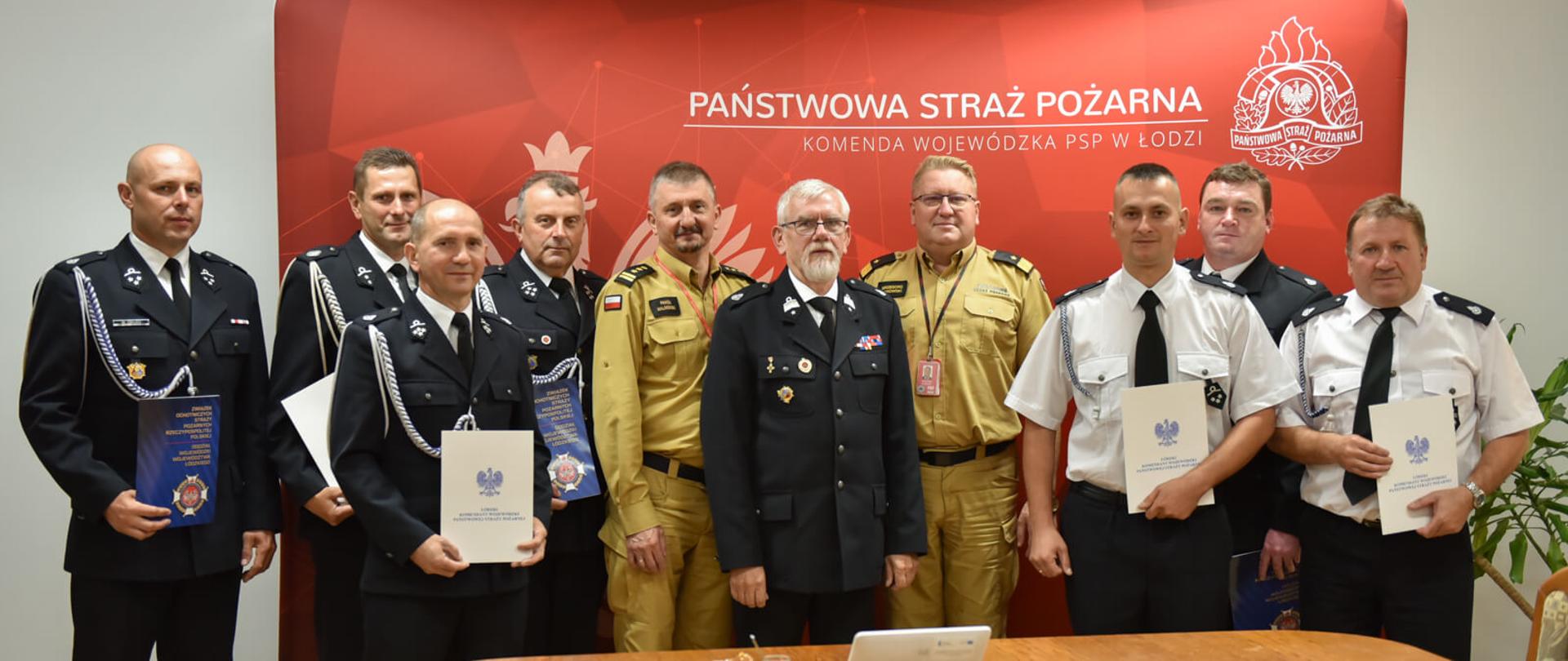 ośmiu druhów i dwóch strażaków pozują do zdjęcia na tle banneru komendy wojewódzkiej PSP w Łodzi