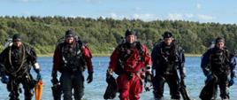 Na zdjęciu widać pięciu nurków w pełnym sprzęcie do nurkowania, wychodzących z wody na tle jeziora i lasu. Ubrani są w suche skafandry i wyposażeni w butle z powietrzem, latarki oraz inne akcesoria niezbędne do działań podwodnych.