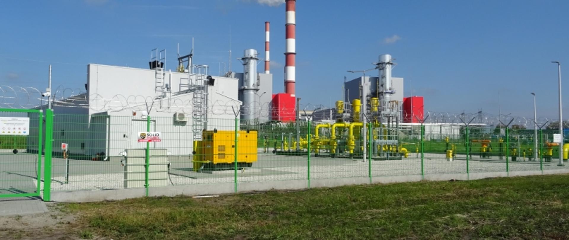 Zakład przemysłowy z ogrodzeniem zabezpieczającym, budynkami, maszynami i kominami w czerwono-białe pasy.