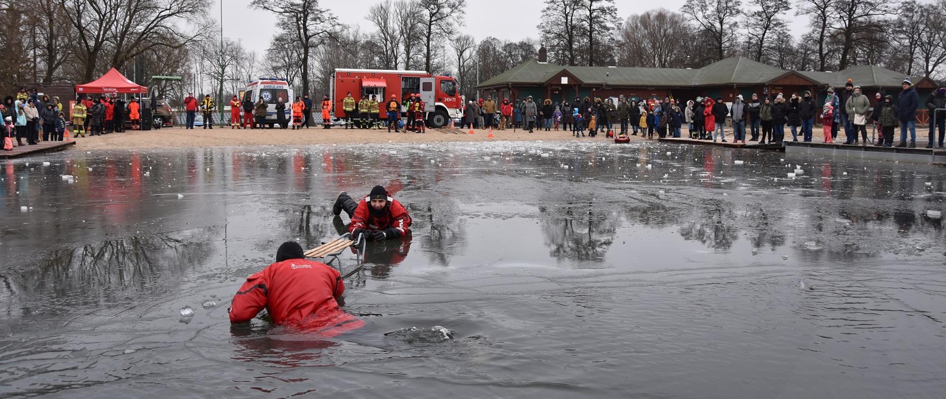 Zdjęcie obrazuje osobę w wodzie a druga będąc na lodzie ją ratuje podając sanki w tle ludzie i samochody