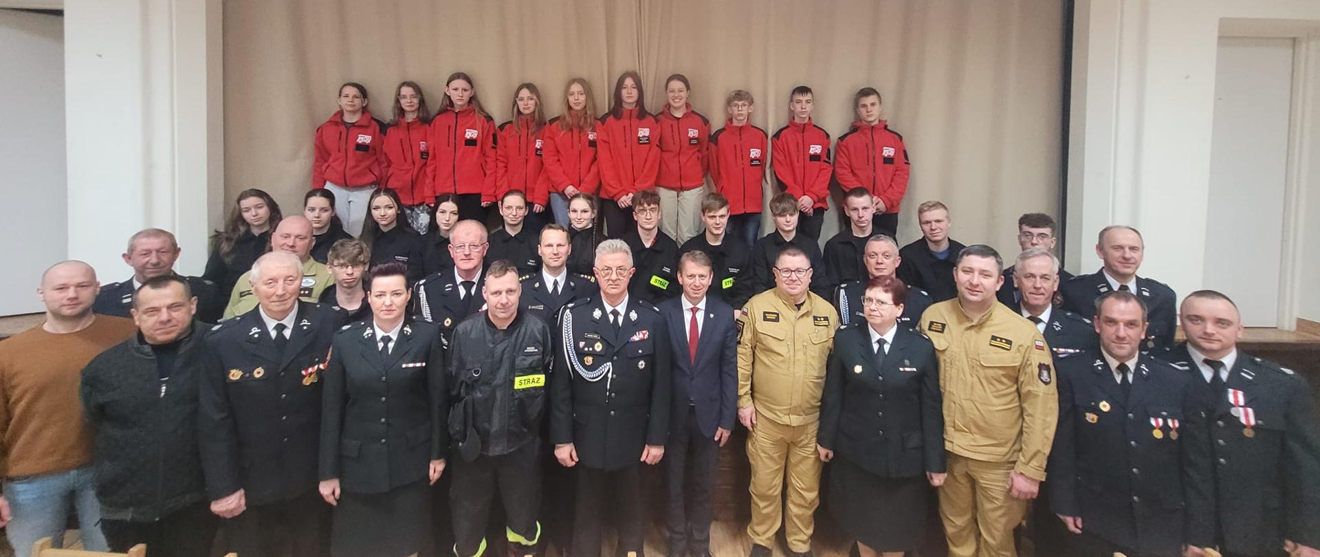 Zdjęcie grupowe uczestników zebrania sprawozdawczego, fotografia wykonana wewnątrz pomieszczenia na tle kurtyny scenicznej. Na zdjęciu grupa około 50 uczestników w trzech rzędach. Wielkość osób z ubrana w mundurach. 
